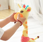 A close-up of a person's hands holding a yellow plush giraffe toy with orange spots and colorful hair tufts in a bright indoor setting.